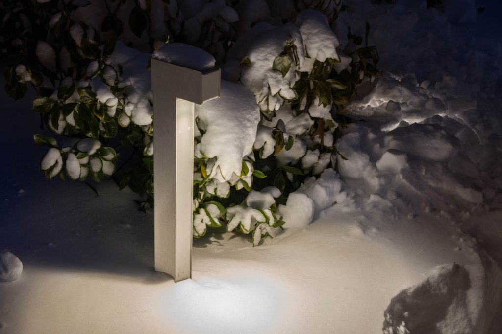 A modern outdoor lamp illuminates snow-covered ground and leafy bushes at night. The lamp and surrounding foliage are dusted with fresh snow, creating a serene winter scene.