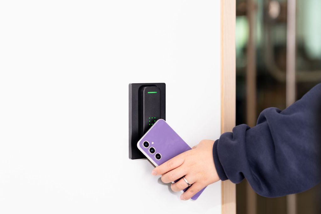 A person holds a purple smartphone up to an electronic card reader on a white wall, likely using the phone for contactless access.