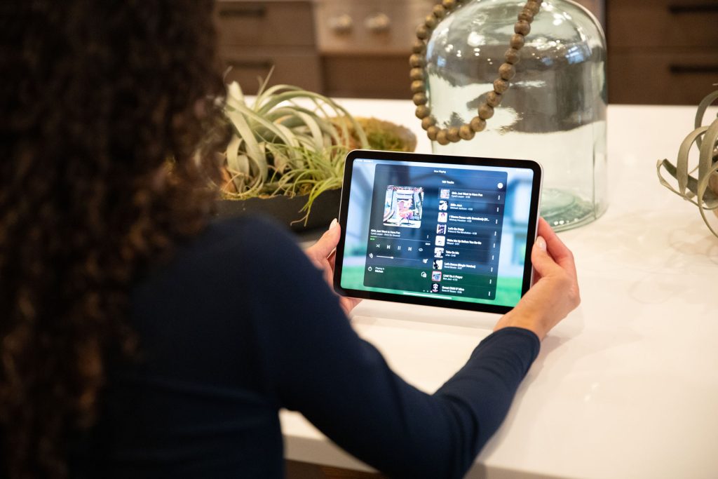 A person with curly hair sits at a kitchen counter holding a tablet, which displays a music playlist. Decorative plants and a large glass jar are on the counter in the background.