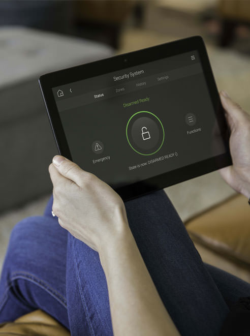 A person holding a tablet displaying a home security system interface, showing the system is disarmed and ready, with options for emergency and functions visible on the screen.
