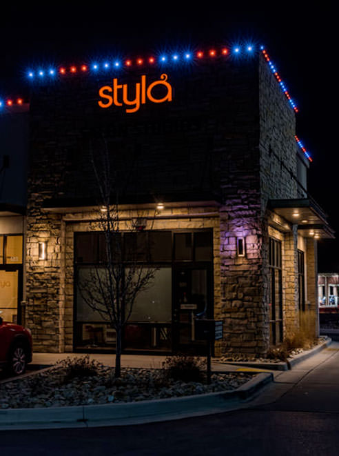A stone building at night with styla in orange letters on top, decorated with blue and red lights along the roof. The entrance is lit, and a bare tree stands in front.